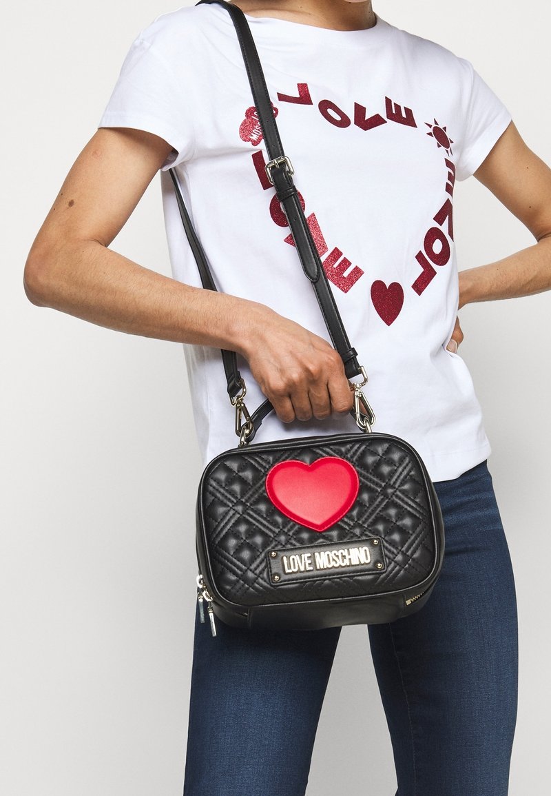 Kvinna i vit t-shirt med röd hjärtformad text och blå jeans som håller en svart vadderad Love Moschino-handväska med röd hjärtdetalj.