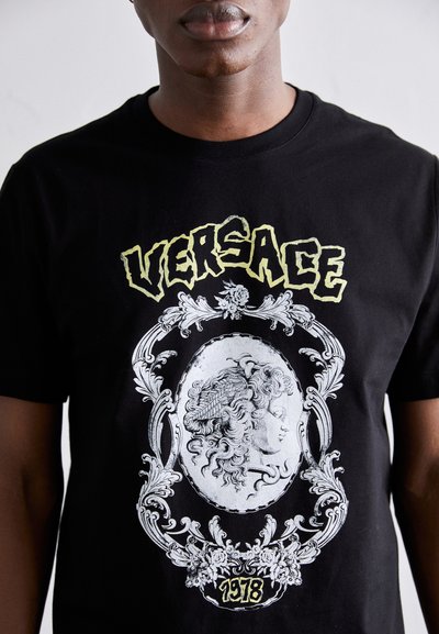Must have must t-särk mustast puuvillast, millel on suur hõbedane klassikalise näo graafika, ornamentele piiratud kujundusega, koos "VERSACE" ja "1978" kollases kirjas.