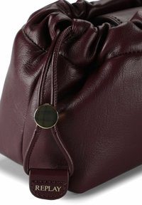 Donker bordeauxleren trekkoordzak met ronde zwarte en gouden sluiting en een "REPLAY" label op de gestikte flap.