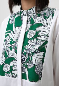 Camicia bianca con un pannello a motivo floreale verde. Presenta un colletto alto e una chiusura a bottoni, con raffinati disegni di foglie in nero.