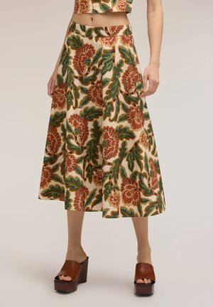 Mujer con falda midi beige con grandes estampados florales rojos y verdes, combinada con sandalias marrones de plataforma.