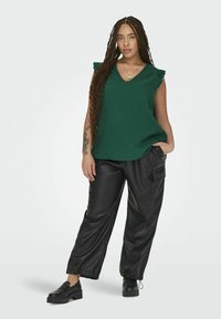 Top sin mangas de textura verde con escote en V, combinado con pantalones anchos de piel sintética negra que presentan bolsillos laterales y puños elásticos. Zapatos negros.