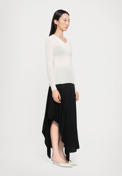 Haut à manches longues en dentelle blanche à côtes associé à une jupe noire asymétrique. La tenue présente un décolleté en V et des textures lisses. Des mules blanches complètent le look.