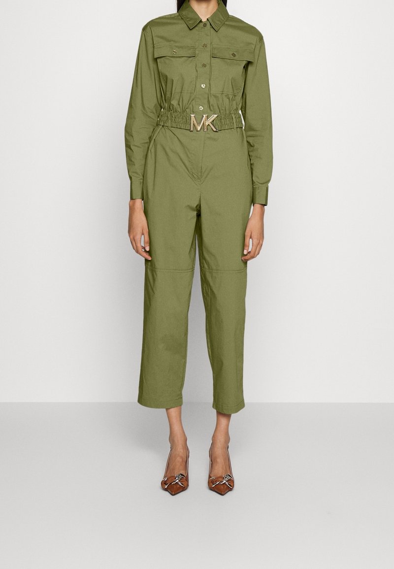 Femme portant une combinaison manches longues vert olive avec poches poitrine, ceinture élastique à boucle MK et escarpins marron à bout pointu.