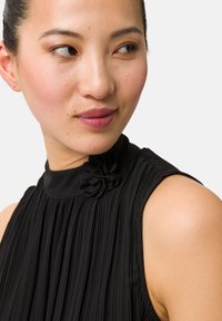 Svart plisserad klänning med hög krage och en blommig accent vid halsen. Slät textur med skräddarsydd passform och ärmlös design.