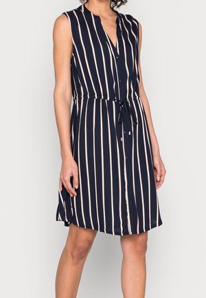 Femme portant une robe sans manches bleu marine à rayures blanches verticales, boutonnée à l'avant, avec une ceinture à la taille, se tenant devant un fond clair uni.