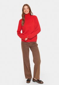 Moteris stovi, dėvinti raudoną megztinį su aukšta apykakle, rudas plačias kelnes ir tamsiai rudus mokasinus, šypsosi, suspaudusi rankas priešais save.