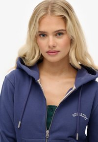 Felpa con zip di colore blu navy, foderata leggera, con cerniera argentata e logo. Presenta un grande cappuccio e un tessuto morbido e strutturato, con tasca a marsupio.