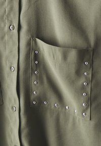 Olive green shirt featuring a pocket with silver studs and button closures along the front. Fabric has a textured finish.