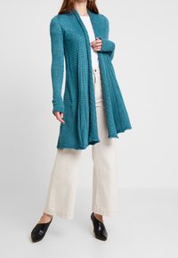 Lång, öppen kappa i teal, ribbad textur, med långa ärmar och tumhål. I kombination med ljusa vida byxor och svarta klackar.