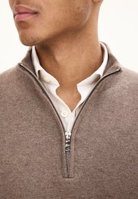 Maglione brunito con zip realizzato in morbido tessuto, con zip argento e una camicia bianca con colletto sotto. Vista ravvicinata del collo.