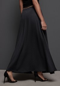 Jupe maxi noire texturée avec une silhouette fluide, taille élastique et motif de vagues subtil. Portée avec des chaussures à talons hauts noires.