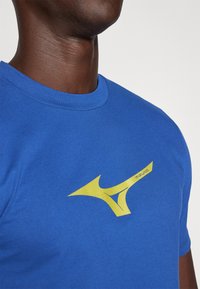 Zils kokvilnas T-krekls ar izteiktu dzeltenu Mizuno logo uz krūškurvja. Ar apaļu kakla dizainu, īsām piedurknēm un gludu tekstūru.