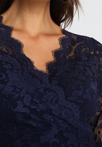 Granatowa koronkowa bluzka z falbaniastym dekoltem w kształcie litery V i długimi rękawami. Zawiera wzory kwiatowe oraz strukturalne wykończenie tkaniny.
