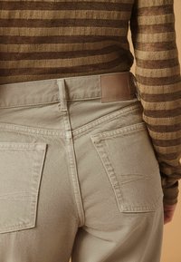 Pantalons beiges en tissu texturé, dotés de deux poches arrière bien visibles et d'une pièce en cuir étiquetée "DENIM.82" sur la taille.