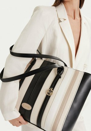 Mujer con un blazer blanco sosteniendo un bolso rayado en negro, blanco y beige con un logo dorado de Lasocki y correas de cuero negro.