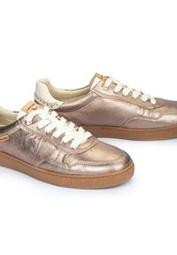 Zapatillas de deporte metálicas en bronce con una textura suave, cordones blancos y un cuello acolchado beige. Presentan una suela de goma con patrón y un detalle de marca.