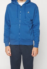 Sudadera azul con cremallera hecha de una mezcla de algodón, con un bolsillo tipo canguro, capucha ajustable con cordón y un pequeño logo blanco en el pecho.