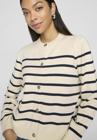 Bēšas krāsas džemperis ar tumši ziliem horizontāliem svītrām, apaļa kakla izgriezums un pieci dekoratīvie pogas. Izceļas ar mīkstu tekstūru un garām piedurknēm.