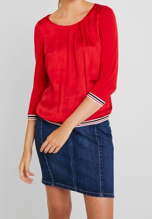 Femme portant un chemisier rouge à manches longues avec des poignets et un ourlet rayés, associé à une jupe en denim bleu sur un fond uni.