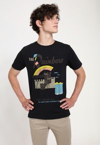 Svart t-shirt med en färgglad grafik av en regnbåge och ett slott med vatten nedan. Texten inkluderar "Regnbågen till Europa" och "Pan American."