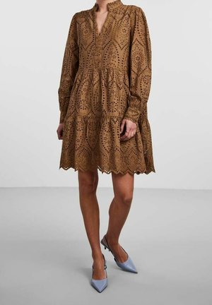 Kvinde iført en brun, langærmet knælængde kjole med hulmønstre og blå slingback stiletter med spids tå.