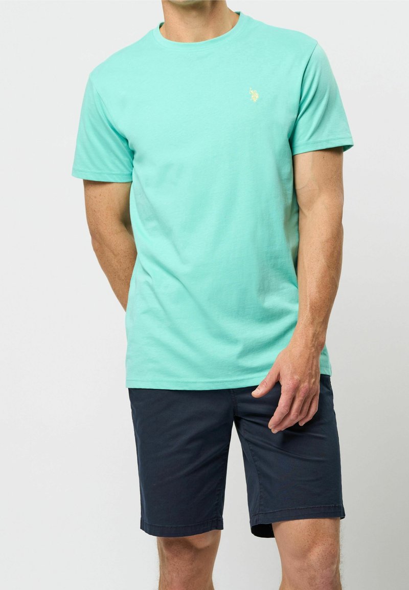 Helles türkisfarbenes Baumwoll-T-Shirt mit rundem Ausschnitt und kurzen Ärmeln, versehen mit einem kleinen goldenen Emblem. Kombiniert mit dunkelblauen Shorts.