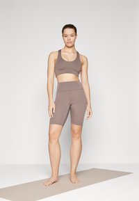 Un reggiseno sportivo marrone e shorts da bicicletta a vita alta abbinati, realizzati in tessuto elasticizzato. La modella è in piedi a piedi nudi su un tappetino da yoga di colore chiaro.