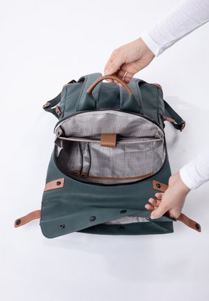 Hände öffnen einen dunkelgrünen Rucksack mit braunen Akzenten und zeigen das leere karierte Innenfutter sowie mehrere Fächer.