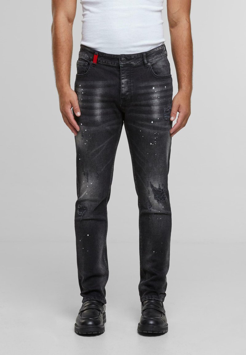 Mann med svarte, slitte skinny jeans med hvite malingssprut, kombinert med svarte lær-sko og en hvit ermeløs skjorte.