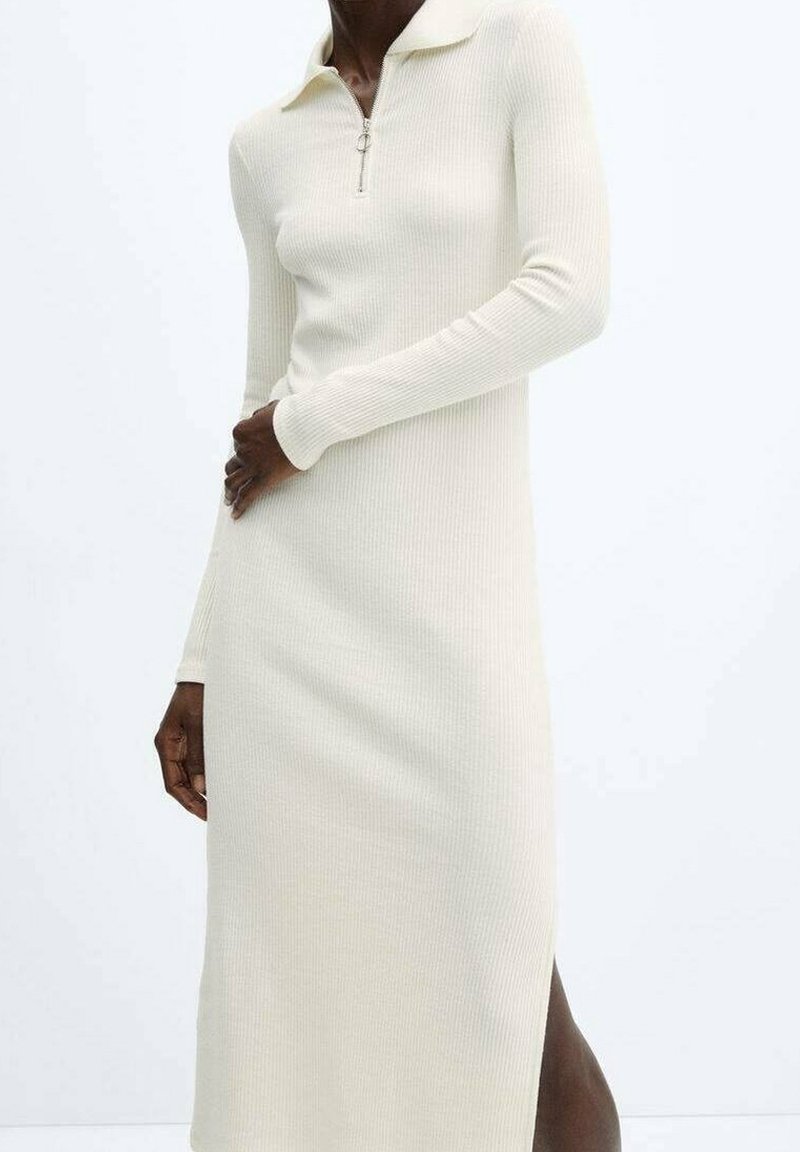Femme portant une robe longue à manches longues en maille côtelée crème avec un col zippé et une fente latérale, debout devant un fond blanc uni.