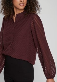 Blouse bordeaux avec un motif en chevrons texturé, fermeture à boutons et longues manches transparentes. Tissu léger avec une coupe ample.