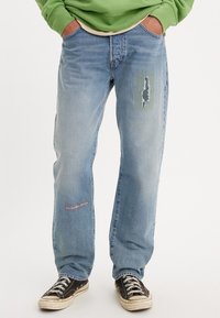 Ljusblå, avslappnade jeans med slitna detaljer, inklusive lappar och sömmar på framsidan. Bärs med svarta sneakers.