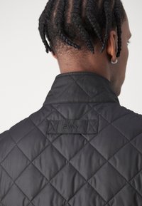 Casaco acolchoado preto com colarinho alto, apresentando um rótulo Barbour bordado na parte de trás. O tecido tem uma textura suave e brilhante.