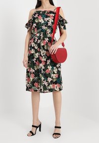 Květinové šaty s odhalenými rameny na černém pozadí, růžové a zelené květiny, s volánovými rukávy. Kombinované s červenou kulatou kabelkou přes rameno a černými lodičkami.
