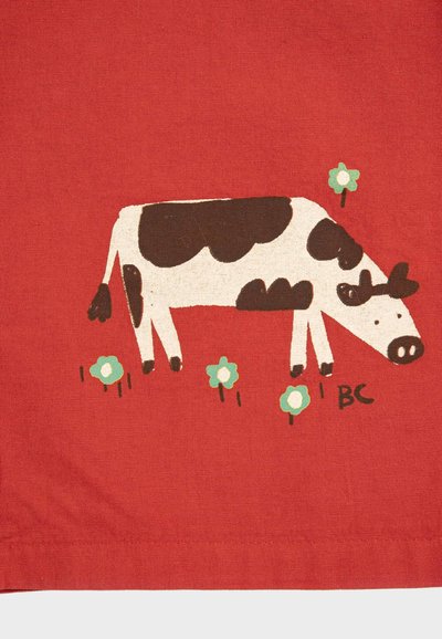 Vache à taches brunes broutant parmi de petites fleurs blanches et vertes sur un fond de tissu rouge texturé.