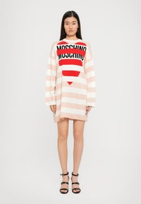 Abito lungo oversized a righe in rosa pallido e bianco, con logo nero e grafica di cuore rosso. Maniche lunghe e vestibilità rilassata.