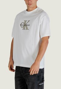 Biały bawełniany t-shirt z krótkimi rękawami, z dużym szaro-czarnym logo Calvin Klein na środku, luźny krój.