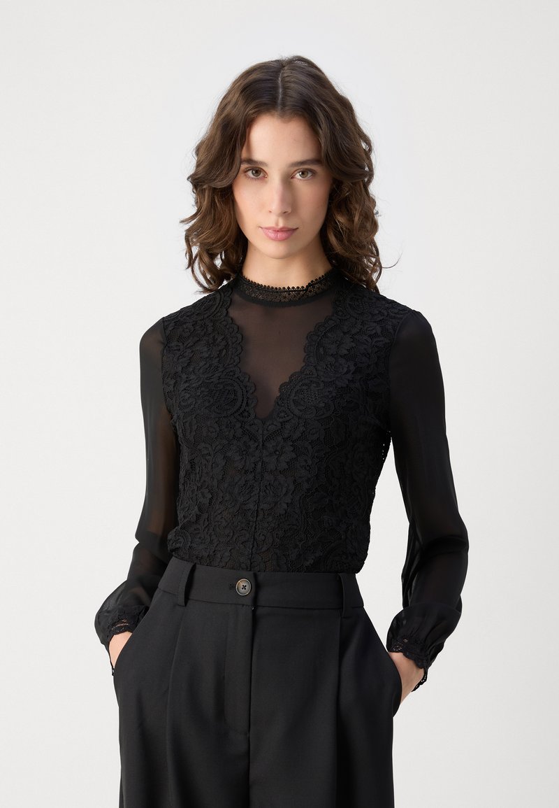 Blusa negra con detalles de encaje, mangas largas transparentes y escote en V. Presenta un delicado borde de encaje en el escote y los puños, combinada con pantalones negros ajustados.