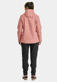 Waterproof pink jacket with a hood, elastic cuffs, and a relaxed fit, paired with black pants and multi-colored trail shoes.