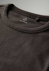 Schwarzes geripptes Strick-T-Shirt mit Rundhalsausschnitt, mit Etikett "THE SET" und "MADE IN BANGLADESH." Glatte Textur, Nahaufnahme.