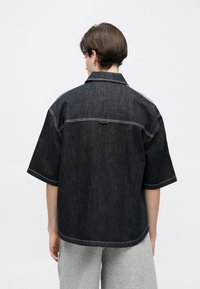 Άτομο με κοντά καστανά μαλλιά που φορά χαλαρό σκούρο τζιν πουκάμισο με κοντά μανίκια και ανοιχτό γκρι παντελόνι, με την πλάτη προς τα εμπρός σε απλό φόντο.