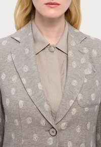 Beež jakk ringikujuliste valgete mustritega, lõhiklapid, ühe nööbiga kinniti ja rinna-tasku. Allpool triibuline särk ümarate kaelustega.