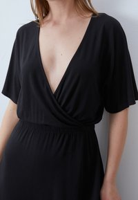 Robe portefeuille noire avec un décolleté en V profond, des manches courtes et une taille élastiquée. Le tissu semble lisse et léger.