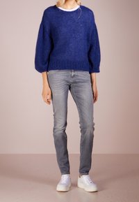 Pull bleu en maille avec manches larges, porté sur une chemise blanche, associé à un jean gris clair et des baskets blanches. Texture lisse, coupe décontractée.