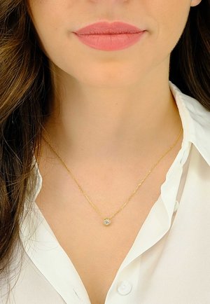Collier chaîne en or avec un pendentif rond en pierre précieuse claire, serti dans un chaton simple, reposant au-dessus d'une chemise blanche à col.