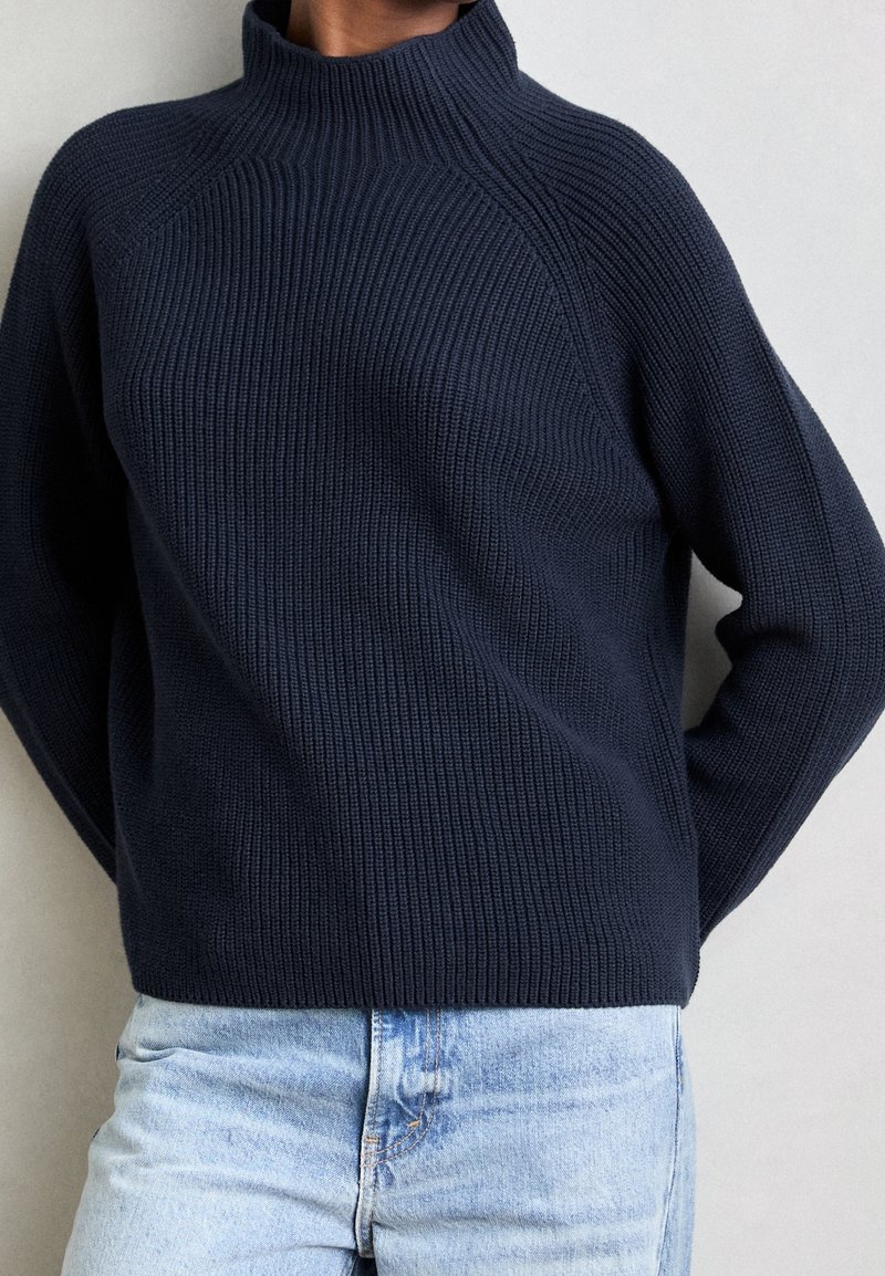 Personne portant un pull en tricot côtelé bleu marine avec un col montant et un jean en denim bleu clair, debout devant un fond uni.