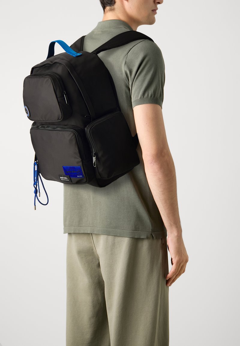 Black backpack with multiple zippered pockets, blue accents, durable fabric, and padded shoulder straps. Worn on the back over a green shirt.