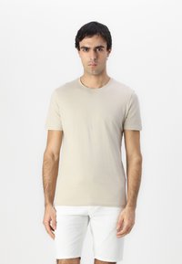 Beige Baumwoll-T-Shirt mit rundem Halsausschnitt und kurzen Ärmeln, präsentiert an einem Modell. Der Stoff wirkt weich und glatt und ist ohne Muster.