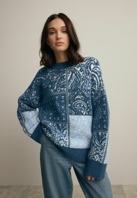Blauer und hellblauer gemusterter Strickpullover mit Rundhalsausschnitt und langen Ärmeln, der ein Patchwork-Design und strukturierte Akzente aufweist.